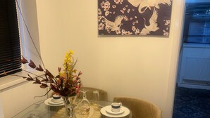 Een eetgedeelte met een glazen tafel gedekt voor vier personen, omringd door beige stoelen. Op de tafel staat een vaas met bloemen, en erboven hangt een schilderij van vogels en bloemen.