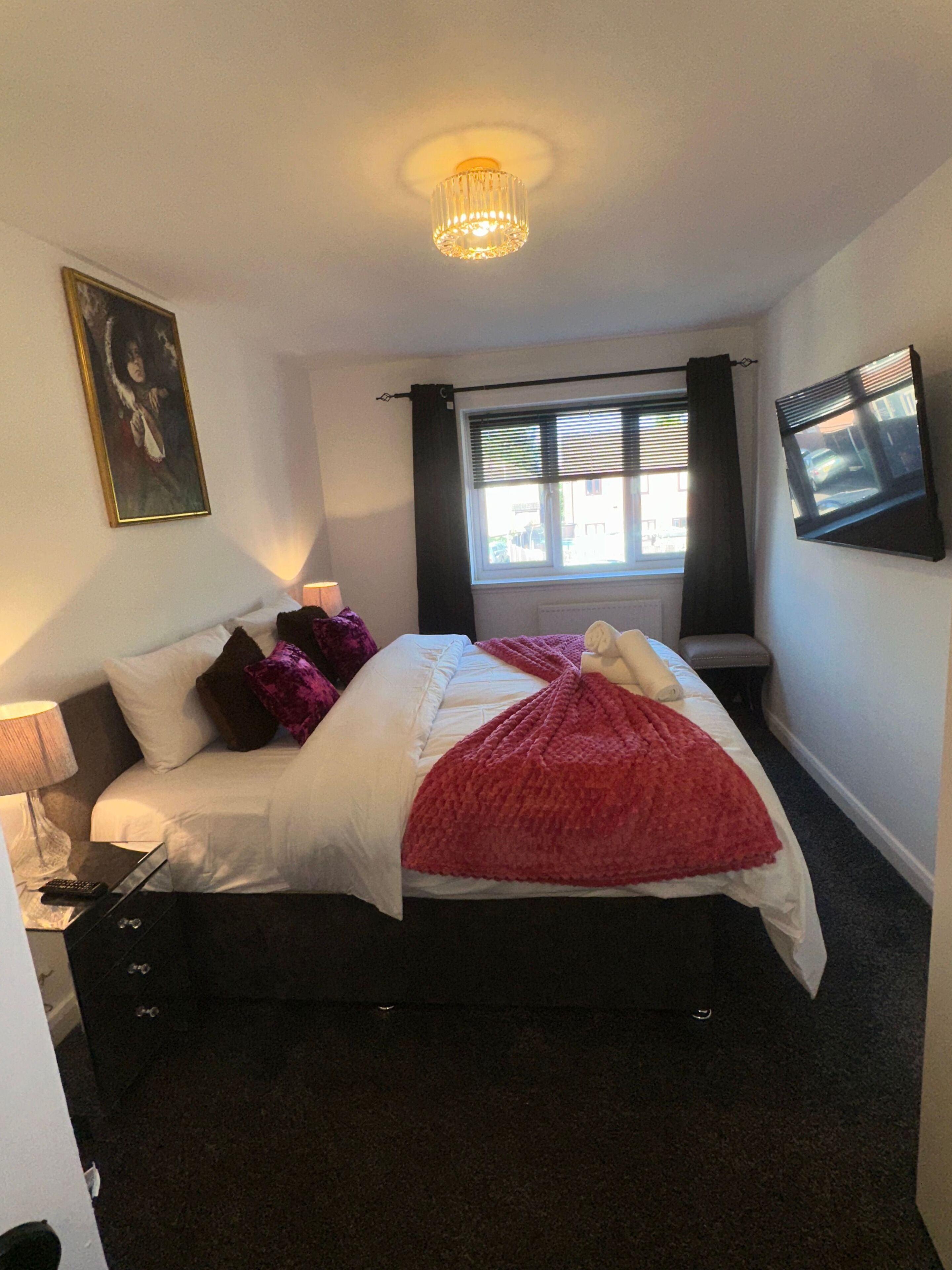 Un dormitorio con cama, lámpara de noche, ventana con cortinas, un cuadro en la pared y televisión montada en la pared.