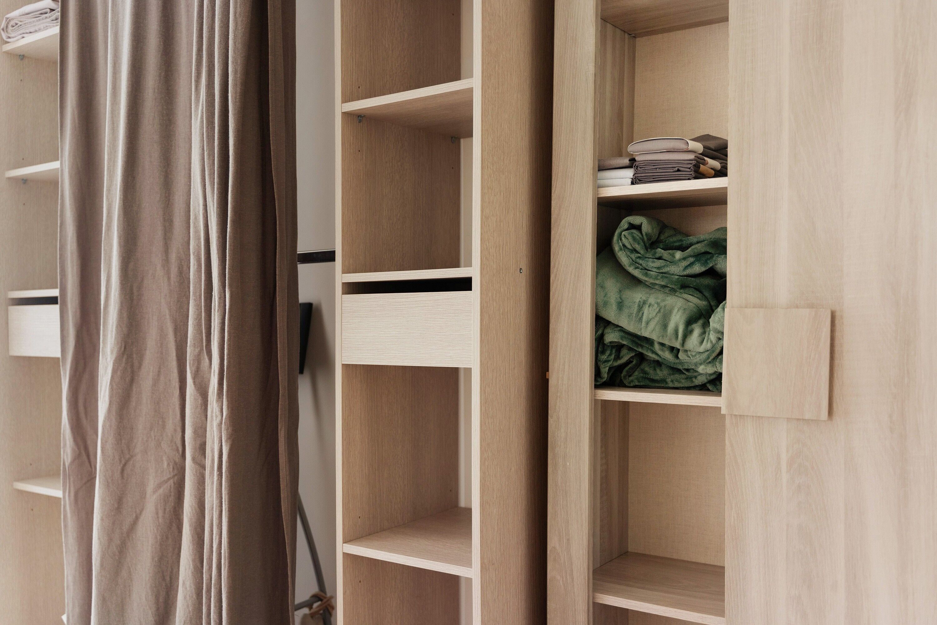 Une armoire en bois, dotée de plusieurs étagères et d’un espace pour suspendre les vêtements, est partiellement ouverte, révélant des vêtements pliés et une couverture verte.