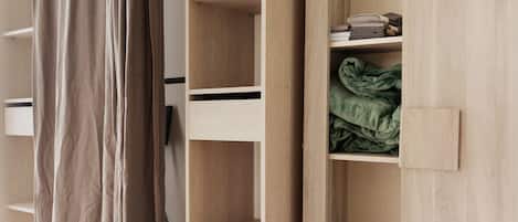 Une armoire en bois, dotée de plusieurs étagères et d’un espace pour suspendre les vêtements, est partiellement ouverte, révélant des vêtements pliés et une couverture verte.