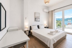 Ein Hotelzimmer mit Bett, Nachttischen, einem Schreibtisch und einem großen Fenster mit Blick auf das Meer und die Hügel.