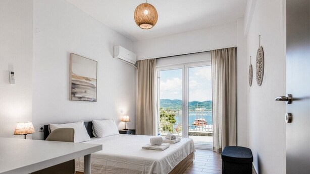 A hotel room with a bed, a desk, a chair, a waste bin, and a view of the sea through a large window. - Phos Residence by Rentals Pro - Neos Marmaras Halkidiki (Neos Marmaras)
