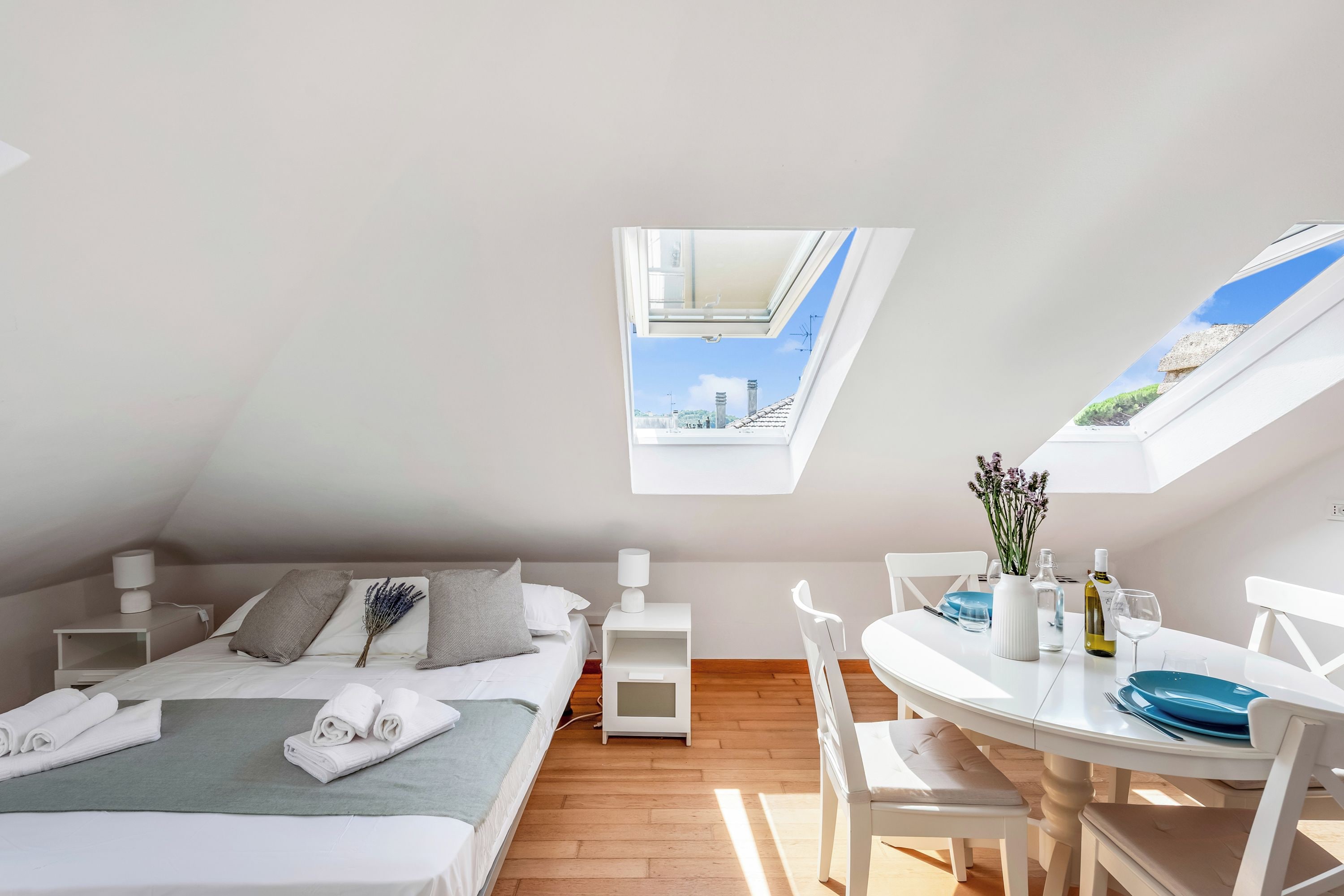 A modern bedroom with a skylight, a bed with pillows, a bedside table with a lamp, and a dining area with a table and chairs.