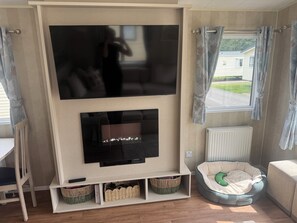 A living room with a flat-screen TV mounted on a wall, a fireplace below, a pet bed, and a window with curtains. - Haggerston Castle Lakeside East 76 (Berwick-upon-Tweed)