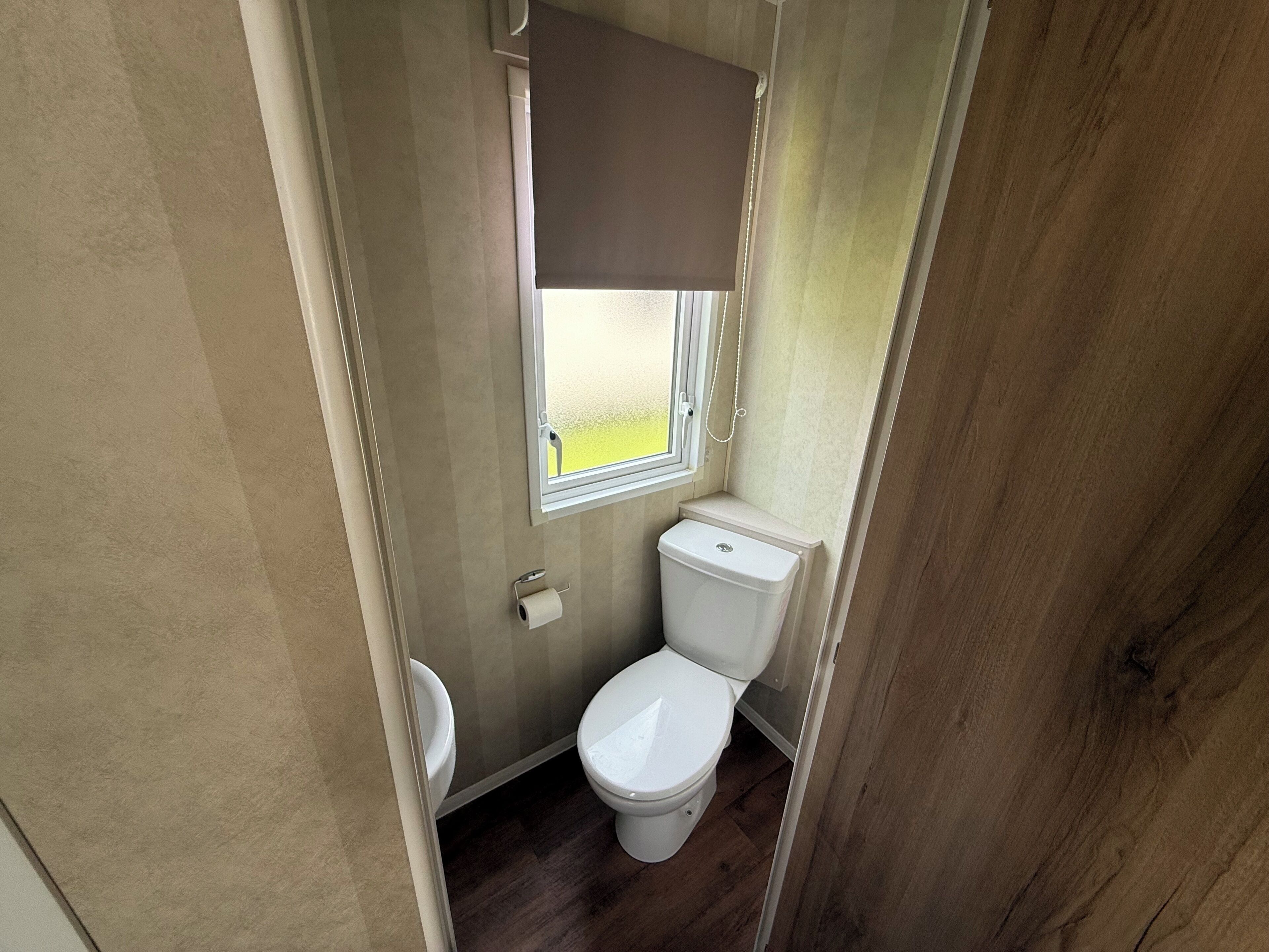 Un baño pequeño con inodoro, una ventana con persiana y un rollo de papel higiénico en la pared.