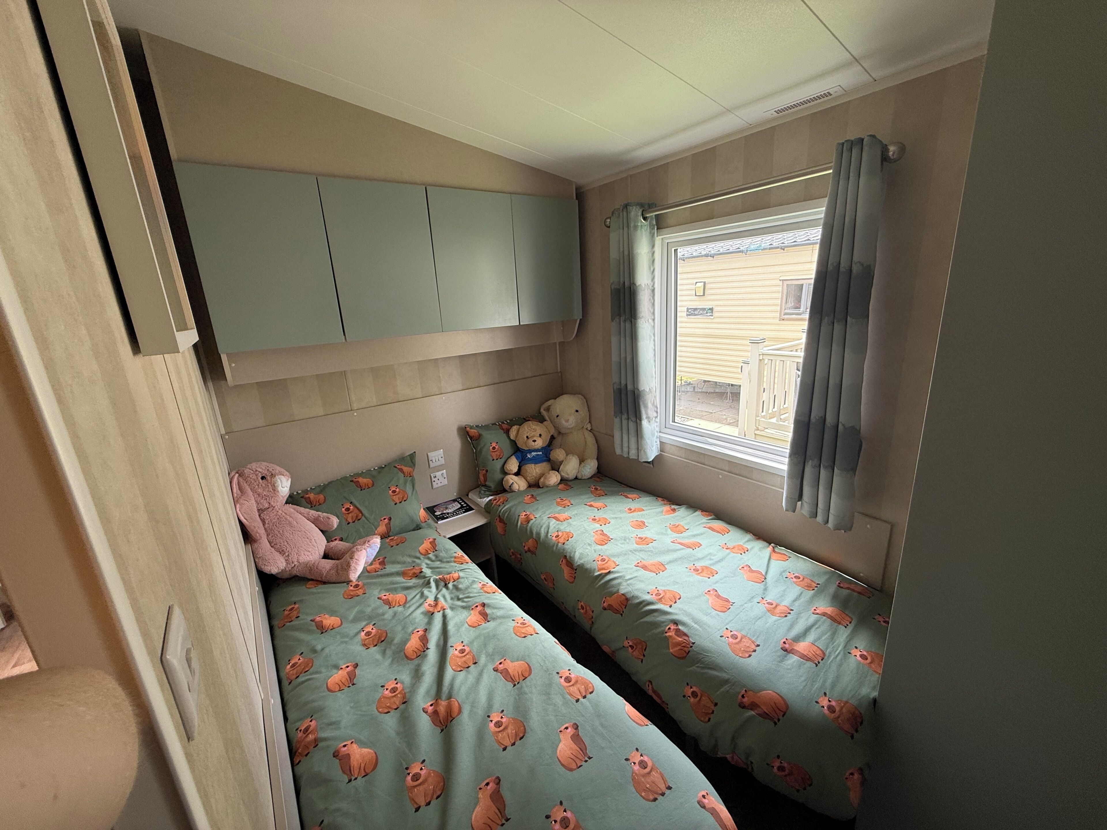 A compact bedroom with two beds, a window with curtains, and a teddy bear on one bed.