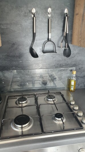A kitchen with a gas stove, three hanging kitchen utensils, and a bottle of oil on the counter. - 2-bedroom seaside condo with mods and cons (Prestatyn)