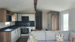 A compact kitchen with modern appliances, a dining area with a grey sofa, and a large window providing natural light. - 2-bedroom seaside condo with mods and cons (Prestatyn)
