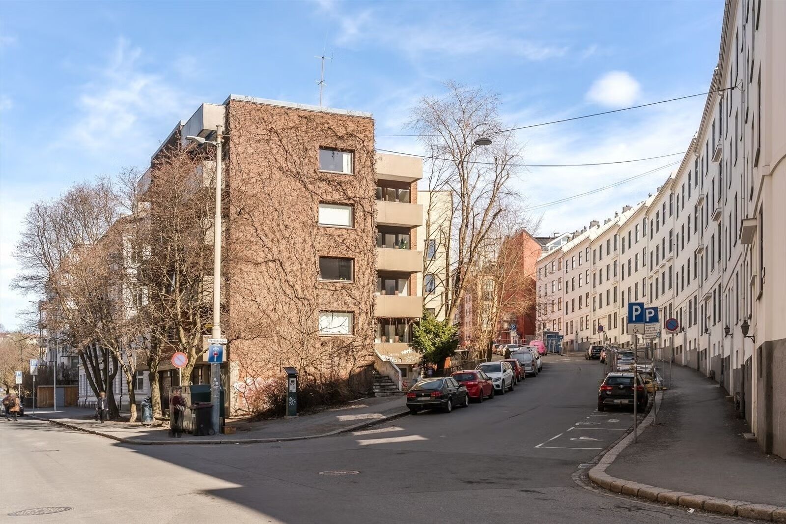 Een straat met geparkeerde auto's, een gebouw met een bakstenen gevel, en een rij witte gebouwen aan de rechterkant.