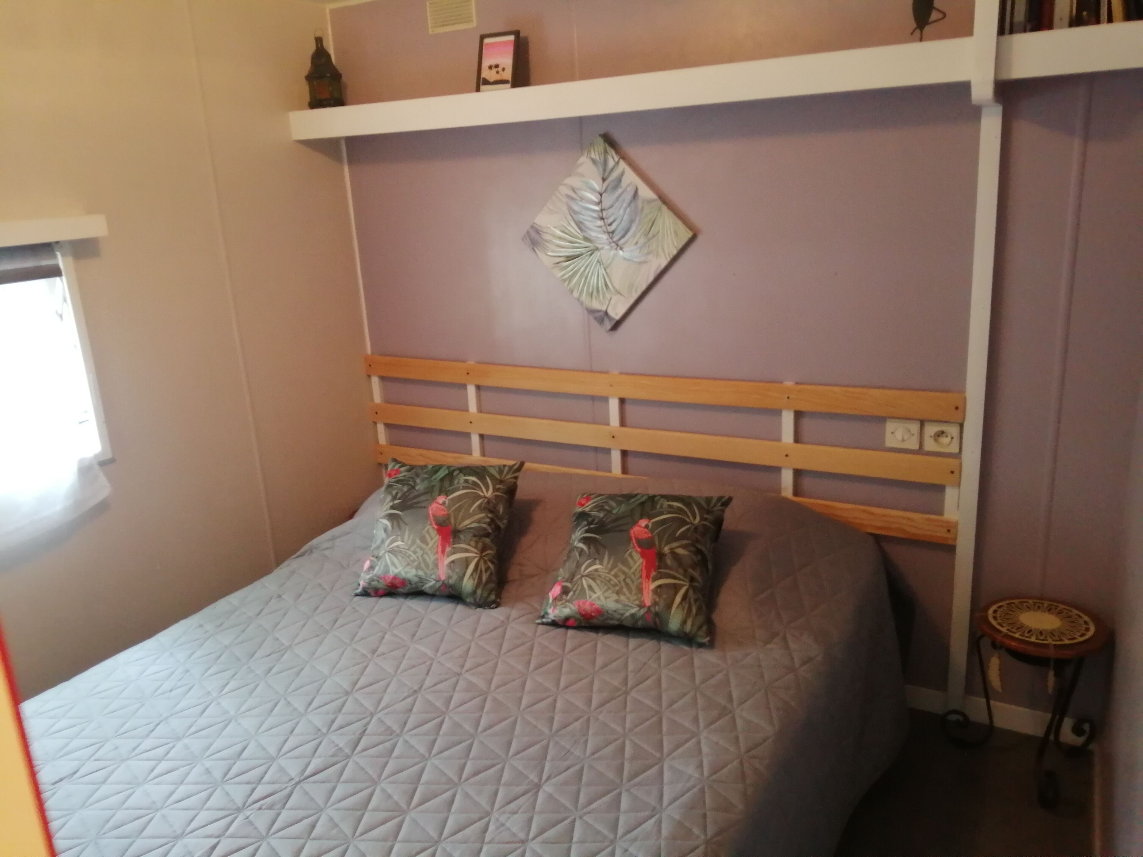 A bed with a wooden headboard, two pillows with a tropical print, and a wall-mounted light fixture.