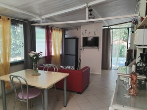 A compact living space with a dining area, a red sofa, a television mounted on the wall, and a kitchen area with a copper pot. - Mobile home in Vias-Plage on private property. (Occitanie)