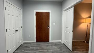 A hallway with a wooden floor, a central door, and a lamp in the adjacent room.