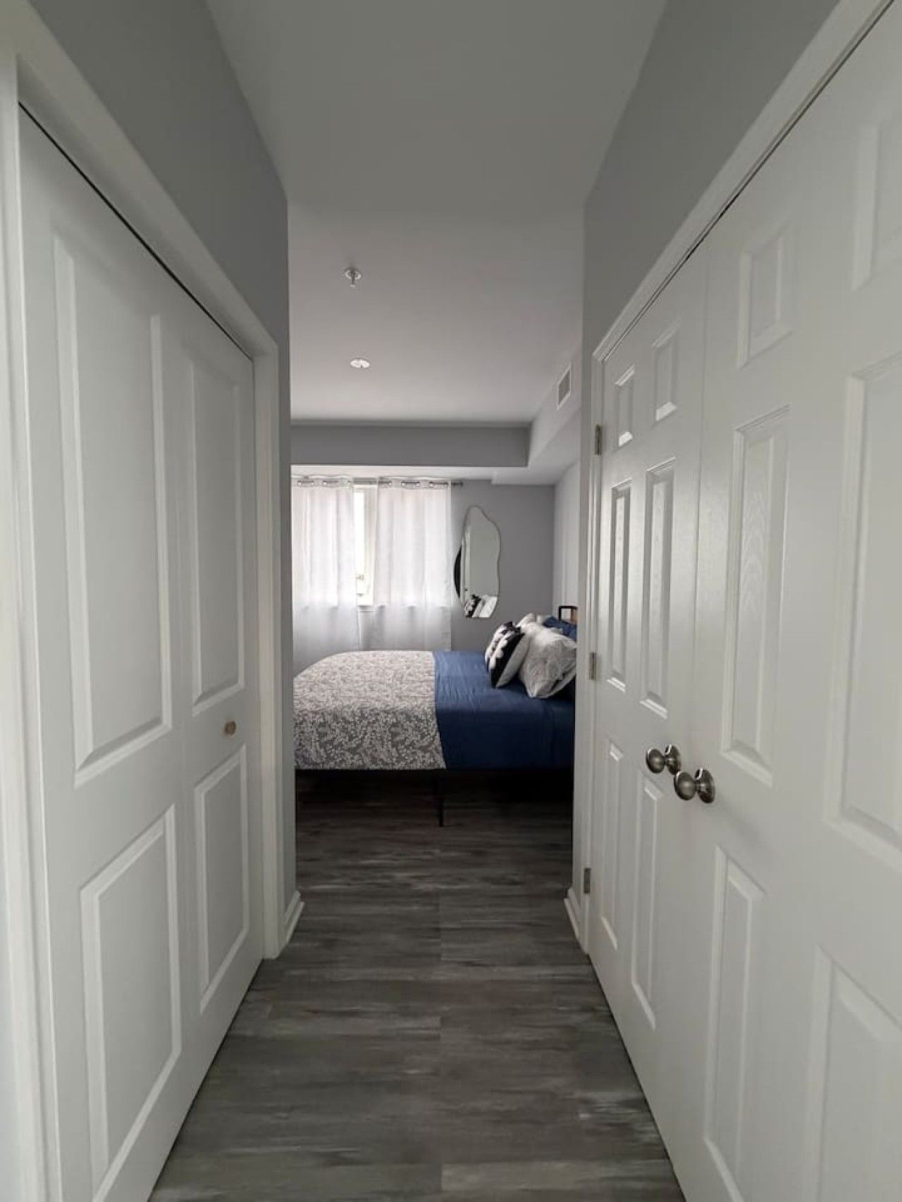A narrow hallway leading to a bedroom with a bed, a mirror, and a wardrobe.