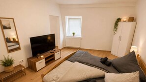 A modern bedroom with a wooden bed, a television, a window with blinds, a cabinet, and a small plant. - Zum Rheinberg - City Studio (Braubach)