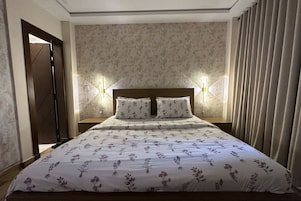 Habitación de hotel con cama, ropa de cama estampada con motivos florales, dos lámparas de noche y una puerta.