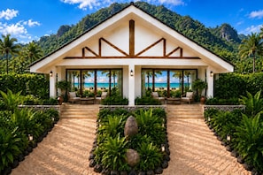 A resort with a covered seating area overlooking a beach, surrounded by tropical vegetation and a mountainous backdrop.