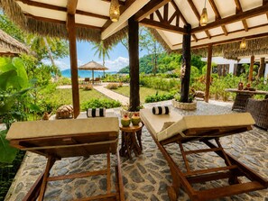 A tropical outdoor lounge area with wooden tables and chairs, a stone floor, and a view of a lush garden and beach.