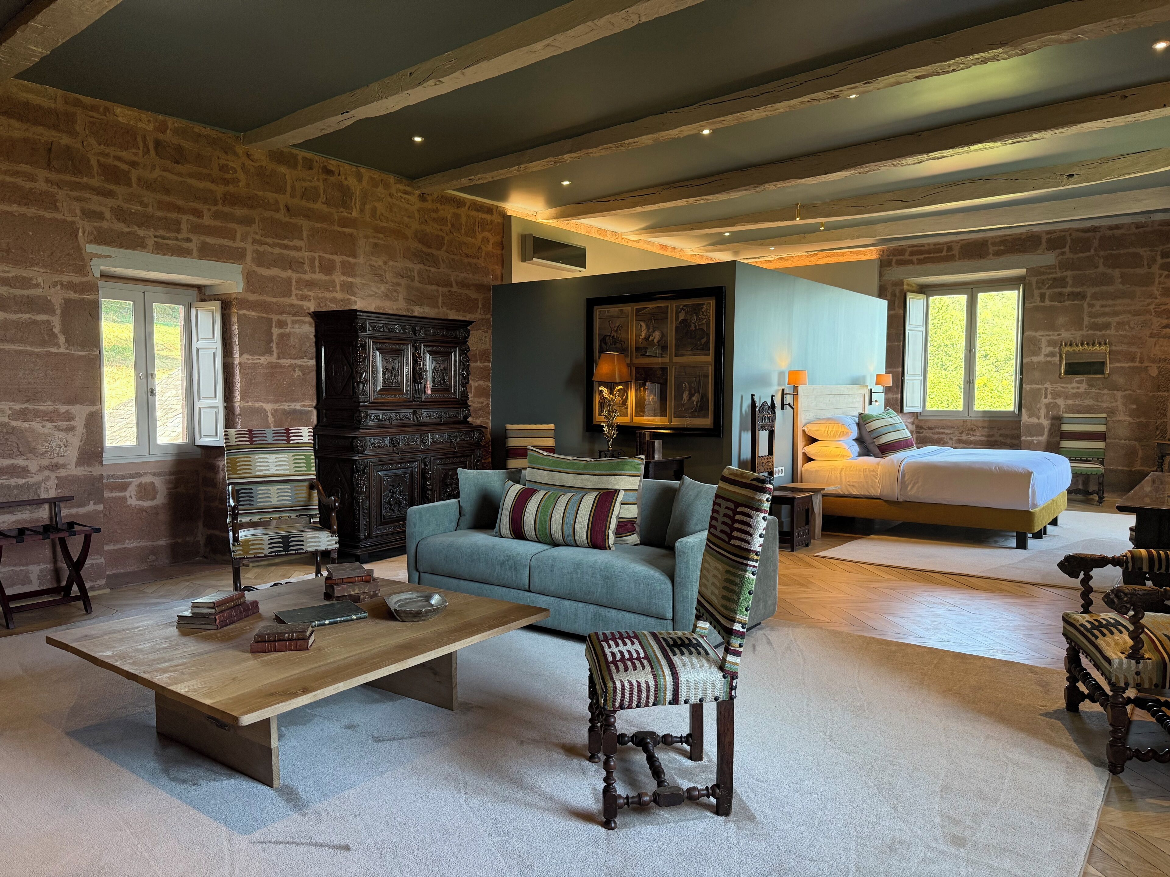 A rustic room with a stone wall, a wooden coffee table, a blue sofa, a wooden chair, a bed with pillows, and a large cabinet.