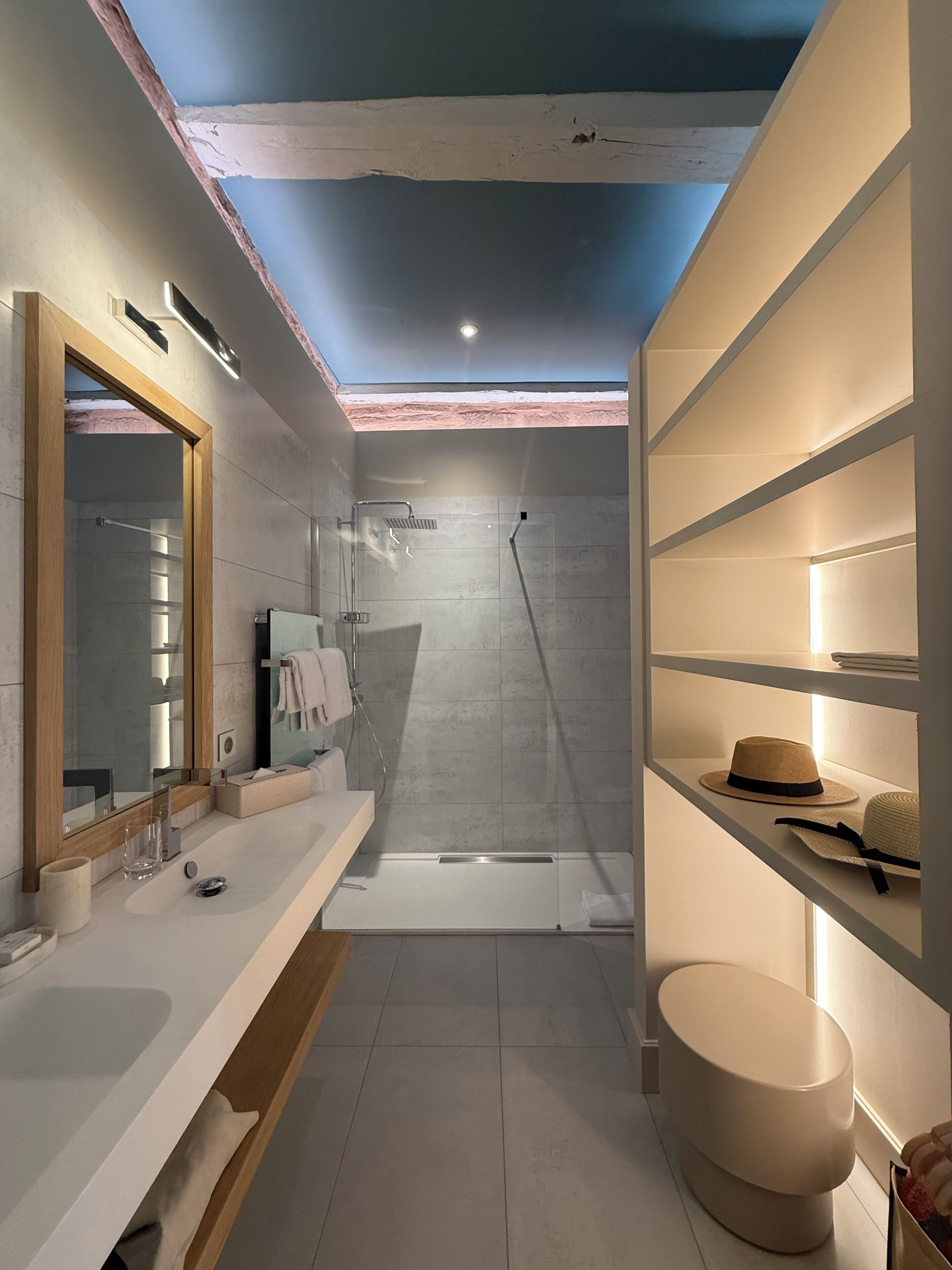 A modern bathroom with a skylight, a shower, a vanity with a mirror, and shelves with hats.