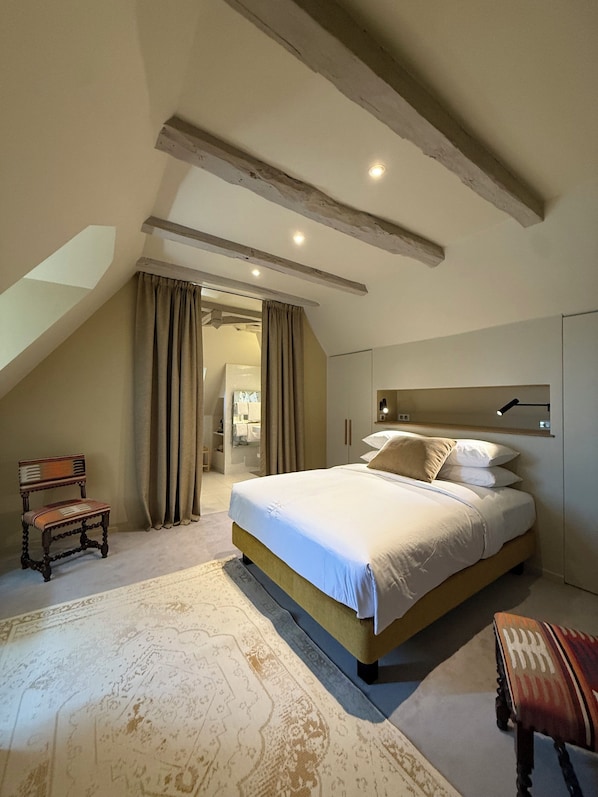 A bedroom with a large bed, a chair, a wardrobe, and a ceiling with exposed beams.