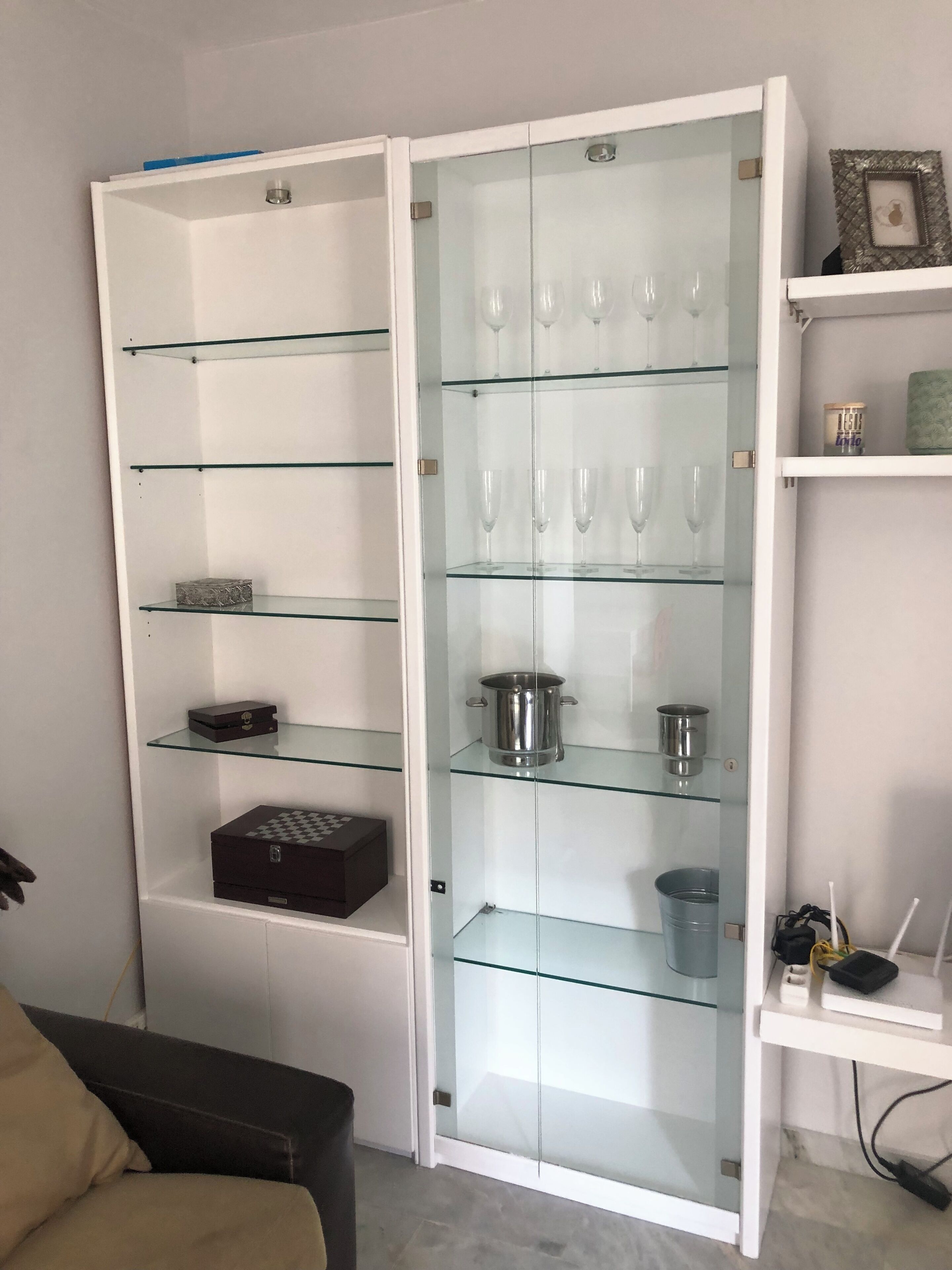 Un módulo de almacenamiento blanco con varias estanterías de vidrio, algunas con objetos como una caja y una olla.