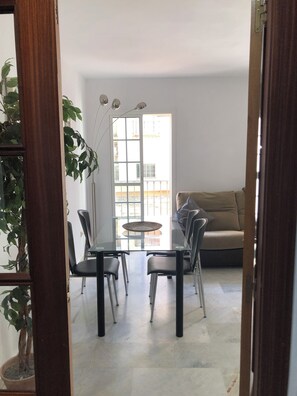 Uma zona de refeições com uma mesa de vidro e cadeiras, um sofá e vista através de uma porta.