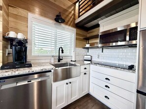 A modern kitchen with stainless steel appliances, white cabinetry, and a window with blinds. - Escalante Escapes Desert Willow Tiny Cabin Rental (Escalante)