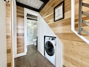 A laundry room with a washing machine, wooden walls, and a door leading to a bathroom. - Escalante Escapes Desert Willow Tiny Cabin Rental (Escalante)