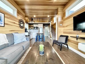 A modern interior with a wooden floor, a sofa, a coffee table with a plant, a TV, and a staircase leading to a kitchen. - Escalante Escapes Desert Willow Tiny Cabin Rental (Escalante)
