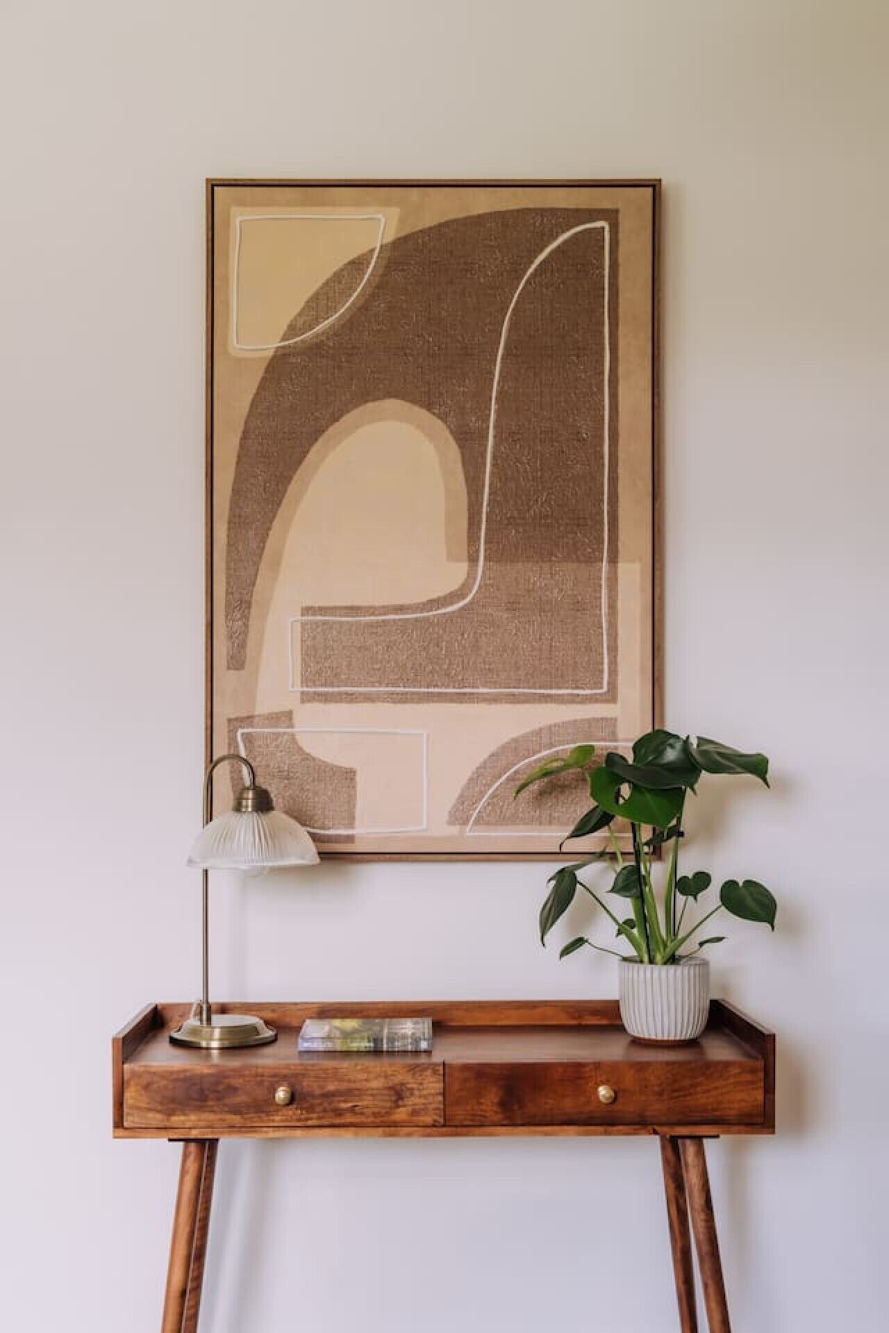 Et indrammet abstrakt kunstværk på en væg, et skrivebord med en lampe og en potteplante ved siden af.