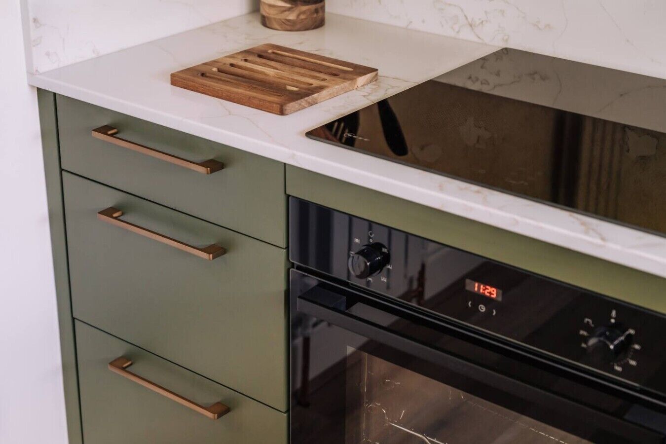 Een keuken met een wit aanrechtblad, een houten snijplank en een klok waarop 11:29 staat.