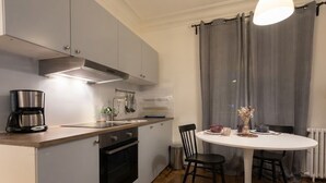 Eine kompakte Küche mit modernen Geräten, ein Essbereich mit weißem Tisch und Stühlen sowie ein Blick durch ein Fenster mit grauen Vorhängen.