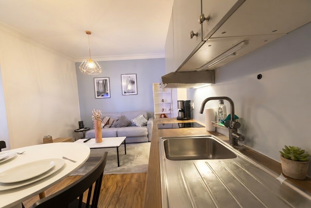 A modern kitchen with a stainless steel sink, a dining area with a table set for two, and a living area with a sofa and a coffee machine.