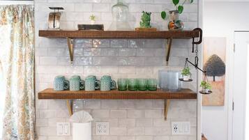 Una cocina con dos estantes de madera que sostienen tazas y cristalería, una planta colgante y un cuadro en la pared.