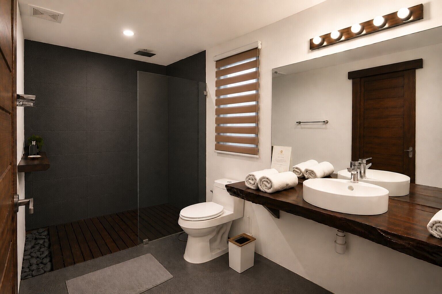 A modern bathroom with a dark tiled shower, a white toilet, and a vanity with two sinks and a mirror.