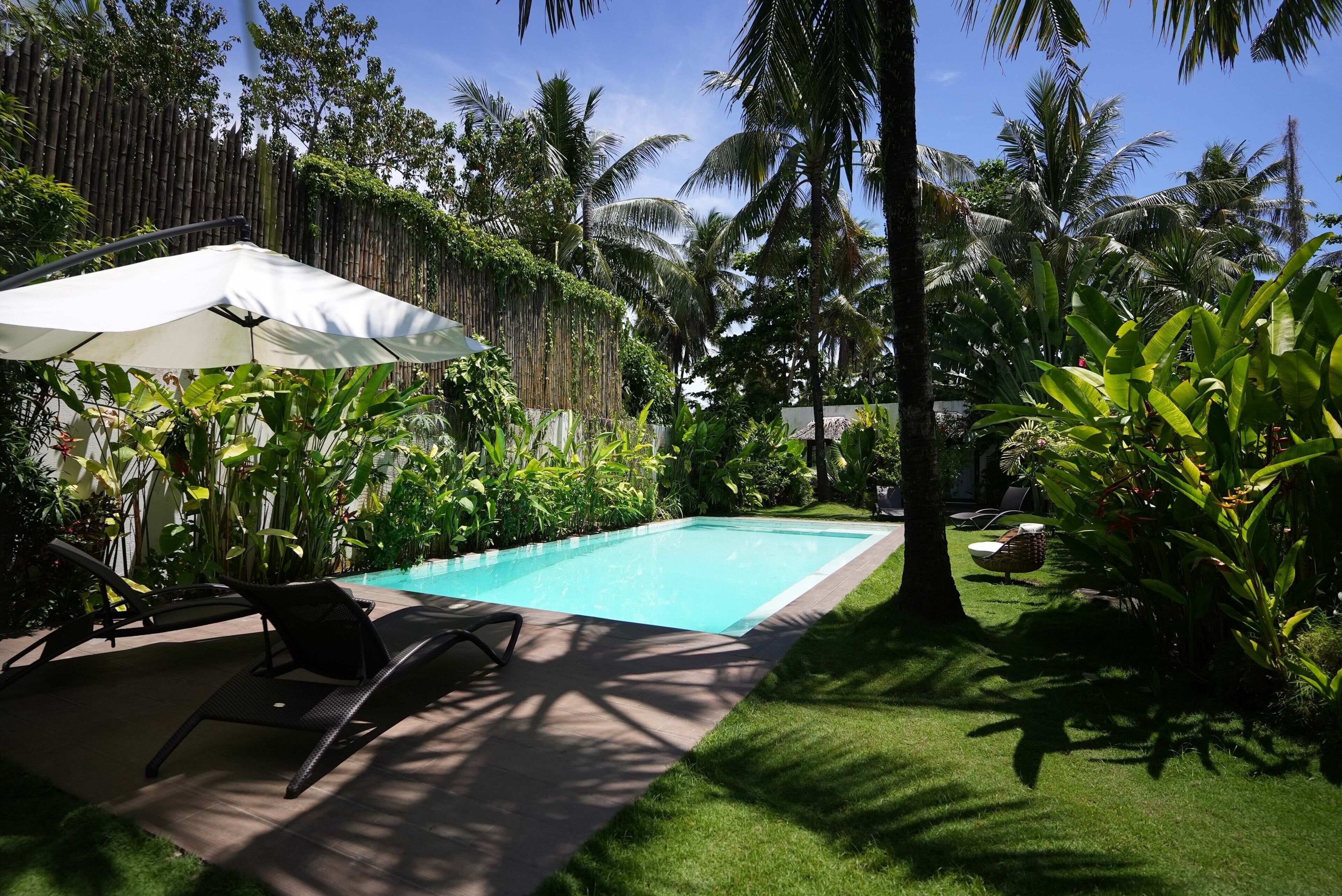 Una piscina con sillones de descanso y una sombrilla, rodeada de exuberante vegetación tropical.