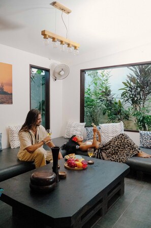 A modern living room with a large window, a dining table with fruit and wine, and a fan. - PALAKA RESORT (General Luna)