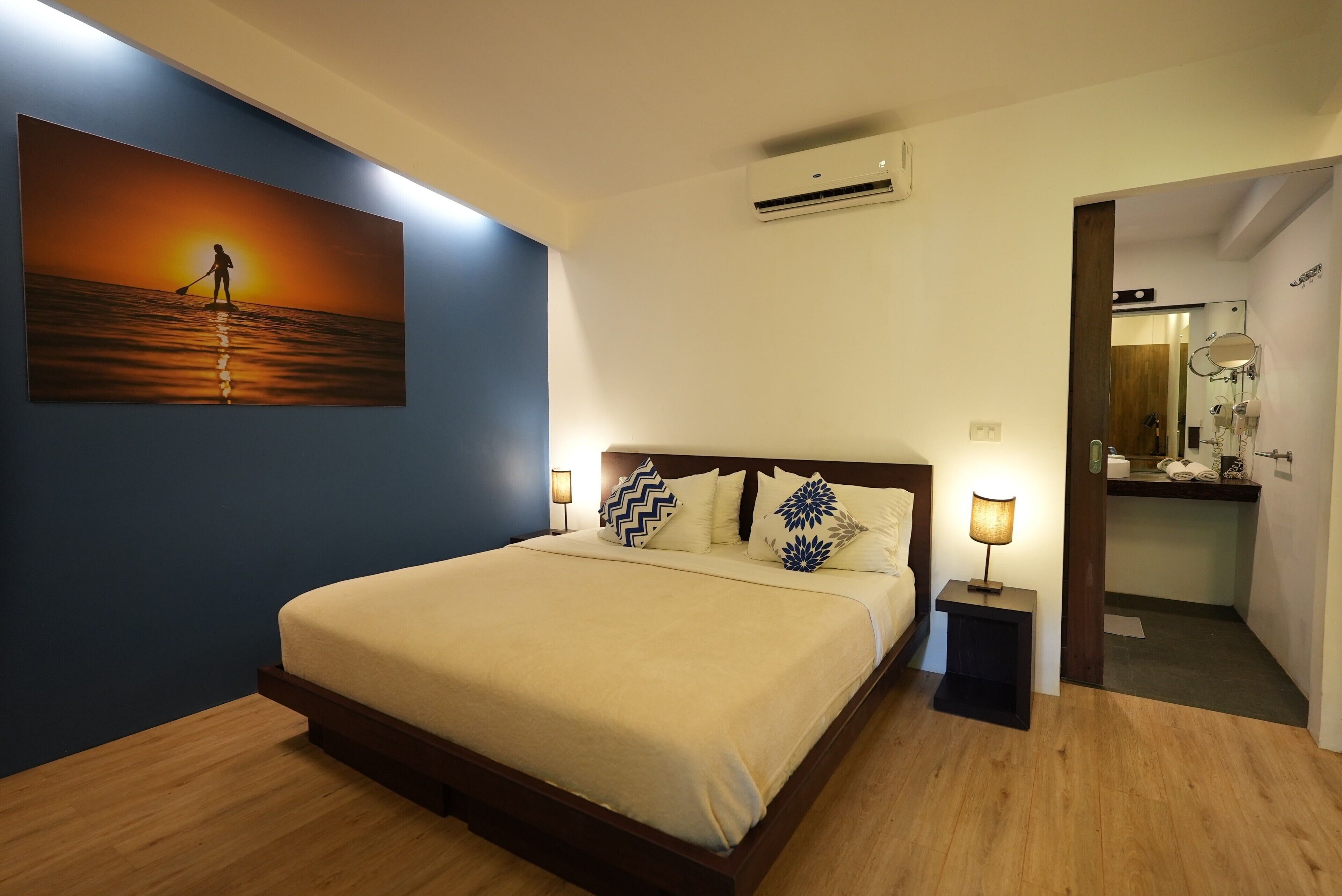 Habitación de hotel con cama, un cuadro de un atardecer, equipo de aire acondicionado, baño con espejo y mesita de noche con lámpara.