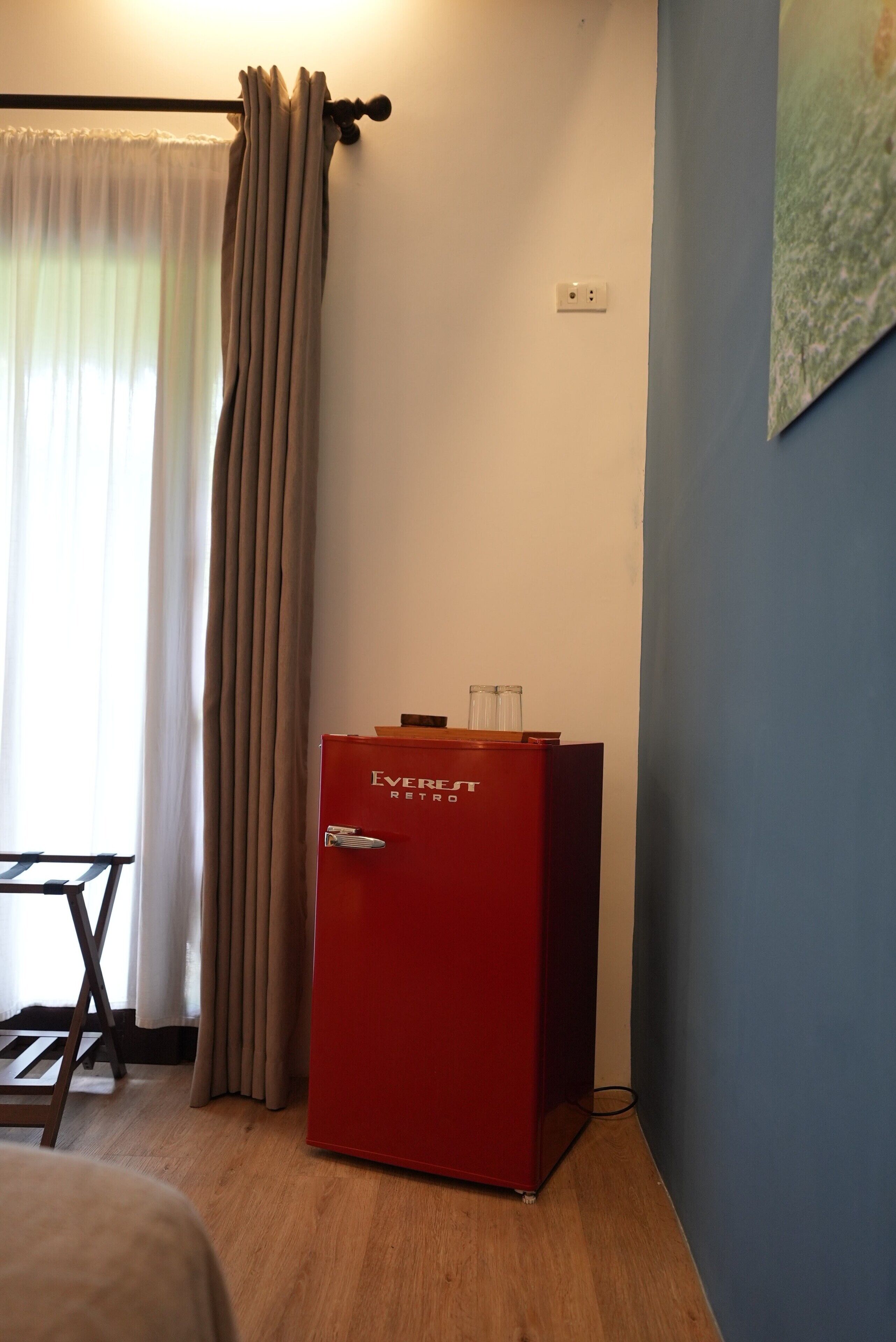 A room with a red minibar, a wooden chair, a window with curtains, and a wall socket.