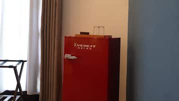Una habitación con minibar rojo, una silla de madera, una ventana con cortinas y un enchufe de pared.