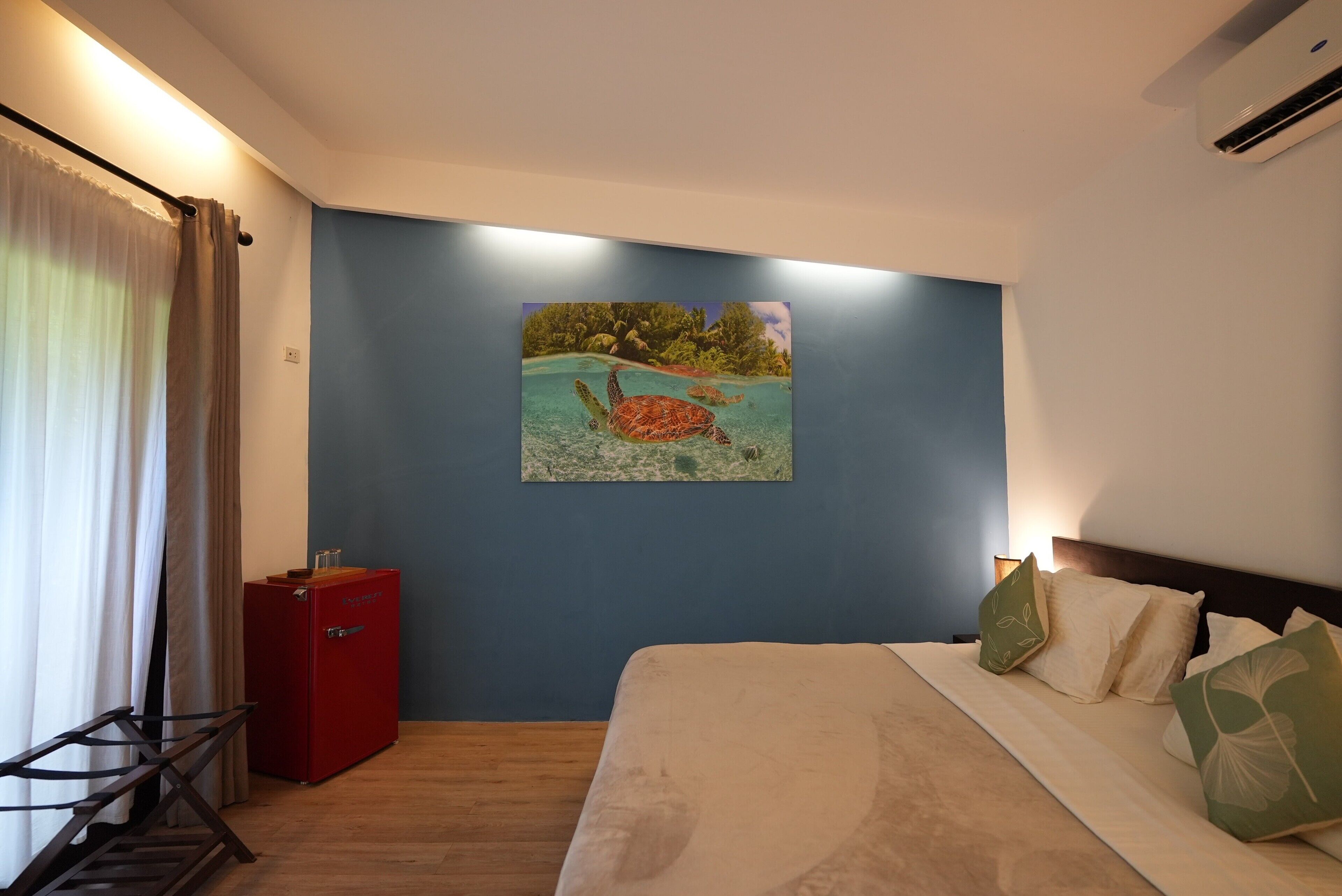Habitación de hotel con cama, un minibar rojo, un cuadro de tortuga marina y una ventana con cortinas.