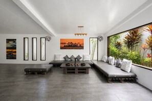 A modern living room with a sofa, a coffee table, and a bench. There are wall-mounted artworks and a large window with a view of plants. - PALAKA RESORT (General Luna)