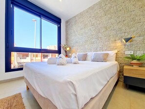 A modern bedroom with a large bed, a stone wall, a view through a window, and a nightstand with a lamp. - Private Roof Terrace & Pool View Penthouse (Mar de Cristal)