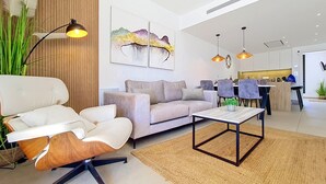 Uma sala de estar moderna com sofá, poltrona e mesa de centro, uma área de jantar com cadeiras e uma cozinha ao fundo.