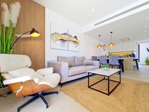 Ein modernes Wohnzimmer mit Sofa, Sessel und Couchtisch, ein Essbereich mit Stühlen und eine Küche im Hintergrund.