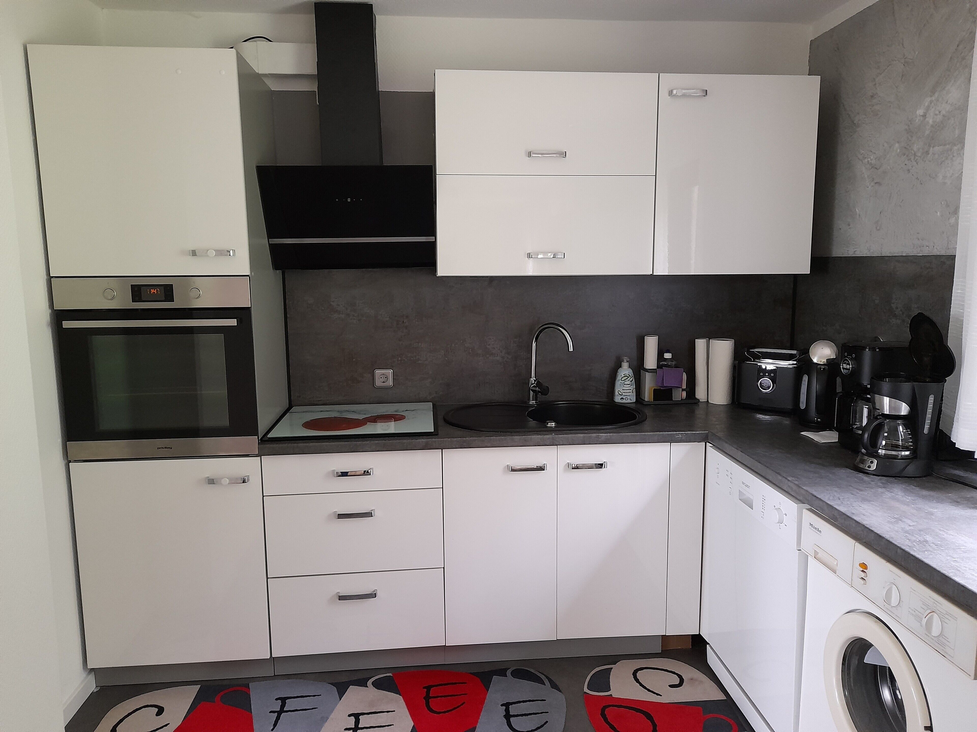 A compact kitchen with white cabinets, a built-in oven, a black range hood, a sink, and a coffee maker.