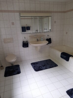 Ein Badezimmer mit Toilette, Waschbecken und Badewanne. Auf dem Boden liegen zwei schwarze Teppiche.