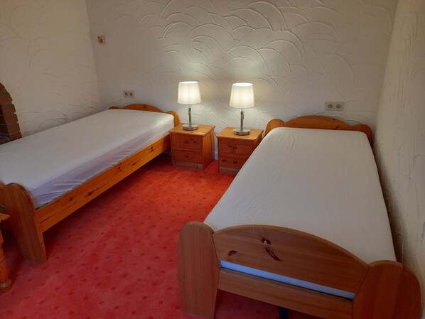 A room with two single beds, a wooden nightstand with lamps, and a red carpet.