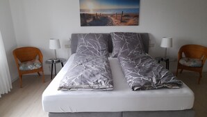 A double bed with grey bedding, two bedside lamps, two wicker chairs, a small table, and a painting of a beach scene on the wall. - Holiday Home 'Am See' with Lake View, Private Terrace and Private Garden (Sundern)