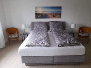 Ein Doppelbett mit grauer Bettwäsche, zwei Nachttischlampen, zwei Korbstühle, ein kleiner Tisch und ein Bild von einer Strandansicht an der Wand.